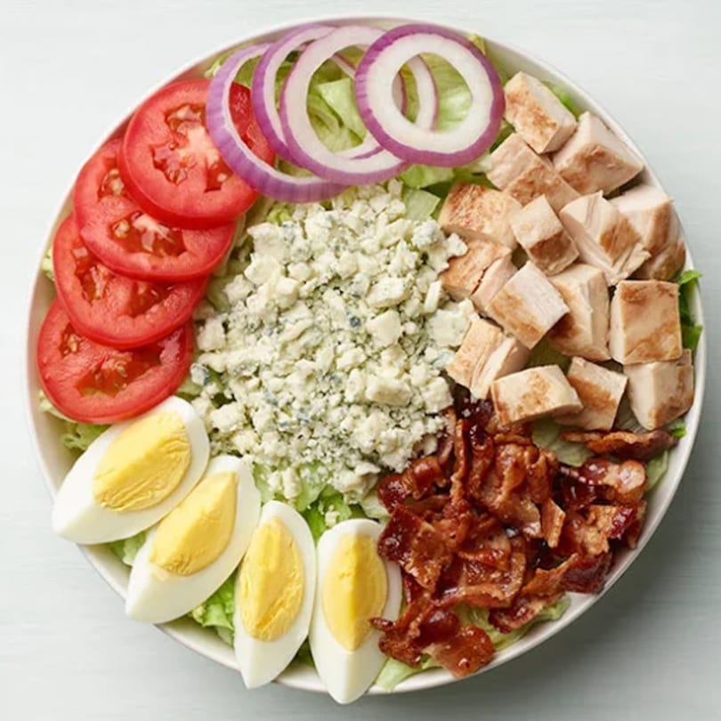 A vibrant Paisanos Pizza salad with lettuce, tomatoes, red onions, blue cheese, chicken, eggs, and bacon.